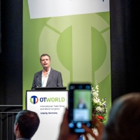 A speaker presents at the OTWorld Congress in Leipzig, while an attendee captures the scene with a smartphone. A speaker presents at the OTWorld Congress in Leipzig, while an attendee captures the scene with a smartphone.