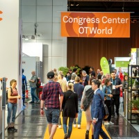 People walk through a trade fair hall with an orange carpet. In the background, a large orange banner reads “Congress Center OTWorld.” On the left side, there is a large advertising display featuring a woman. Attendees move between booths and engage in conversation. People walk through a trade fair hall with an orange carpet. In the background, a large orange banner reads “Congress Center OTWorld.” On the left side, there is a large advertising display featuring a woman. Attendees move between booths and engage in conversation.