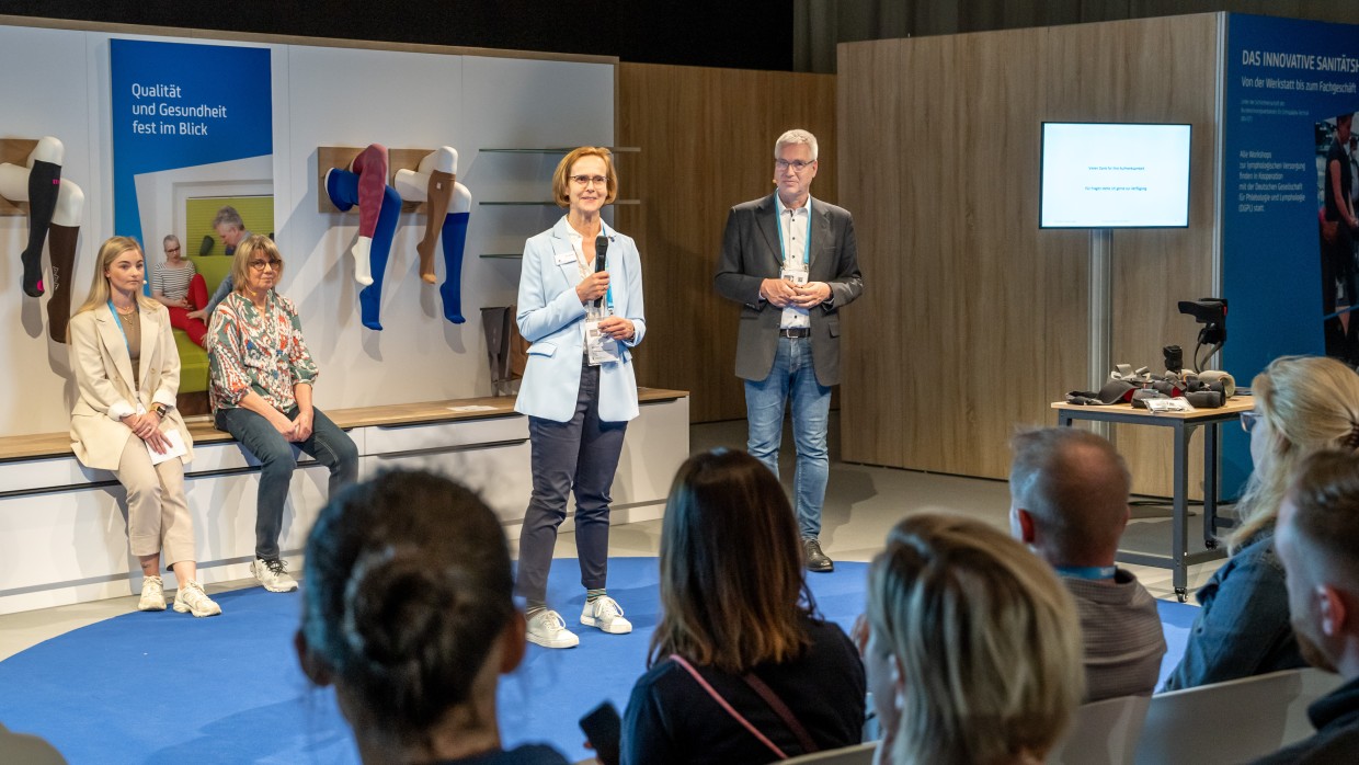 A woman speaks into a microphone on a small stage in front of a seated audience. A man stands next to her, while two other women are seated in the background. Medical compression stockings are displayed on mannequin legs mounted on the wall. A screen and a table with exhibit items are visible on the right. A woman speaks into a microphone on a small stage in front of a seated audience. A man stands next to her, while two other women are seated in the background. Medical compression stockings are displayed on mannequin legs mounted on the wall. A screen and a table with exhibit items are visible on the right.