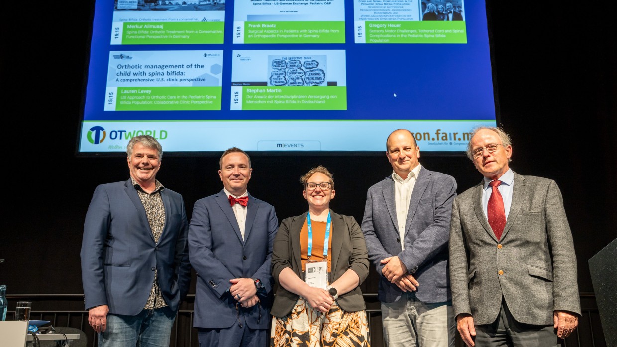 Five people stand side by side on a stage, facing the camera. A large screen behind them displays presentation slides related to orthopedic care. The individuals are wearing conference badges. Five people stand side by side on a stage, facing the camera. A large screen behind them displays presentation slides related to orthopedic care. The individuals are wearing conference badges.
