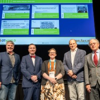 Five people stand side by side on a stage, facing the camera. A large screen behind them displays presentation slides related to orthopedic care. The individuals are wearing conference badges. Five people stand side by side on a stage, facing the camera. A large screen behind them displays presentation slides related to orthopedic care. The individuals are wearing conference badges.
