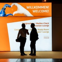 Two people are standing in silhouette in front of a large illuminated welcome screen that reads “WILLKOMMEN! WELCOME!” along with further directions to the congress area. At the top, an illustration shows a human and a bionic hand forming a heart shape. Two people are standing in silhouette in front of a large illuminated welcome screen that reads “WILLKOMMEN! WELCOME!” along with further directions to the congress area. At the top, an illustration shows a human and a bionic hand forming a heart shape.