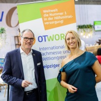 A man in a suit and a woman in a green dress are standing at OTWorld in front of a roll-up banner reading “OTWorld – International Trade Show and World Congress” and the slogan “Number 1 worldwide in assistive technology supply!”; in the background are other trade fair visitors and a lounge area. A man in a suit and a woman in a green dress are standing at OTWorld in front of a roll-up banner reading “OTWorld – International Trade Show and World Congress” and the slogan “Number 1 worldwide in assistive technology supply!”; in the background are other trade fair visitors and a lounge area.