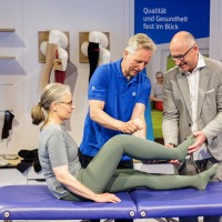 A woman lies on a treatment table at OTWorld while two men – one in a blue polo shirt, the other in a gray suit – hold her legs dressed in green compression garments for a demonstration; in the background, stockings are displayed on wall mounts and a sign reads “Focus on Quality and Health.” A woman lies on a treatment table at OTWorld while two men – one in a blue polo shirt, the other in a gray suit – hold her legs dressed in green compression garments for a demonstration; in the background, stockings are displayed on wall mounts and a sign reads “Focus on Quality and Health.”