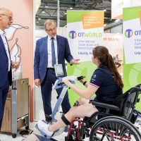 A young woman in a wheelchair uses a pedal-based therapy device at OTWorld, while two men in suits observe the equipment; in the background are two green OTWorld roll-up banners with English and German text. A young woman in a wheelchair uses a pedal-based therapy device at OTWorld, while two men in suits observe the equipment; in the background are two green OTWorld roll-up banners with English and German text.