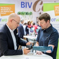 A man in a dark suit examines a blue-black arm prosthesis worn by another man in a dark shirt at OTWorld; in the background are two green OTWorld roll-up banners with English and German text. A man in a dark suit examines a blue-black arm prosthesis worn by another man in a dark shirt at OTWorld; in the background are two green OTWorld roll-up banners with English and German text.